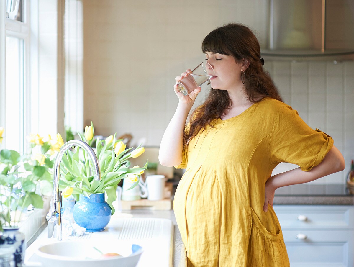 A pregnant woman drinks a glass of water in the kitchen of her home with fresh picked spring flowers in vase.