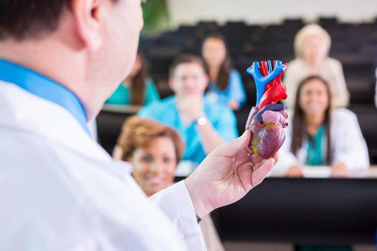 Mature adult Caucasian doctor is speaking to group of heatlhcare professioanls during a medical seminar. He is using an anitomical model of the human heart to explain new technology during his speech. People at medical conference are wearing scrubs and lab coats.