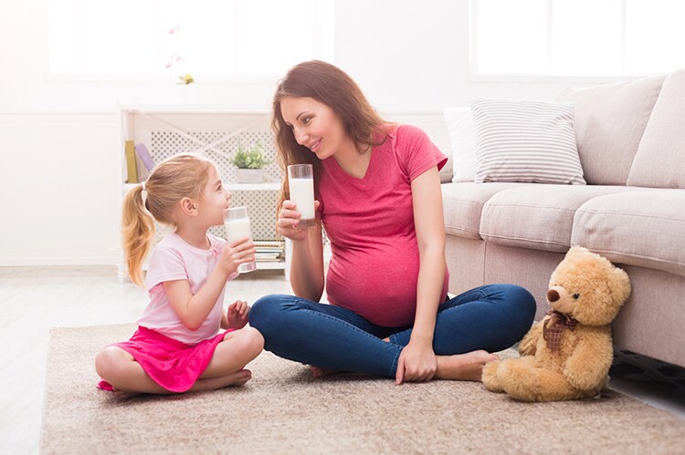 Little girl and her pregnant mom drinking milk sitting on floor at home. Motherhood and care, healthy eating and lifestyle, early development concept, copy space